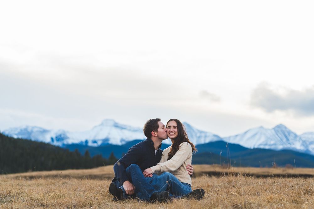 Couple sitting in a golden field with snow-capped mountains, sharing a romantic kiss outdoors.