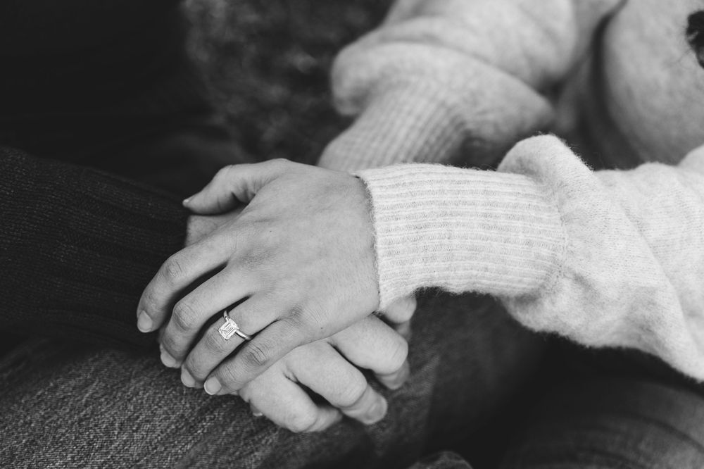 Black and white photo of two people holding hands, one wearing a diamond engagement ring, wearing cozy knit sweaters.