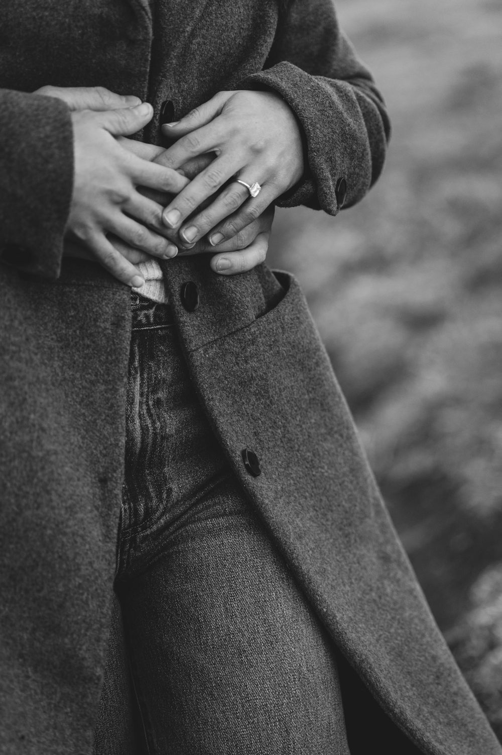 Black and white photo of two hands clasped together, one wearing a ring, over a long wool coat and jeans.