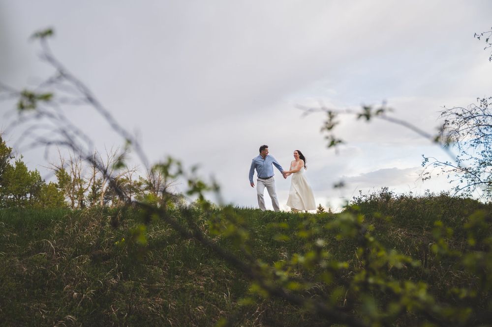 Couple holding hands on a green hill with mountains and cloudy sky in the background during an outdoor engagement session.