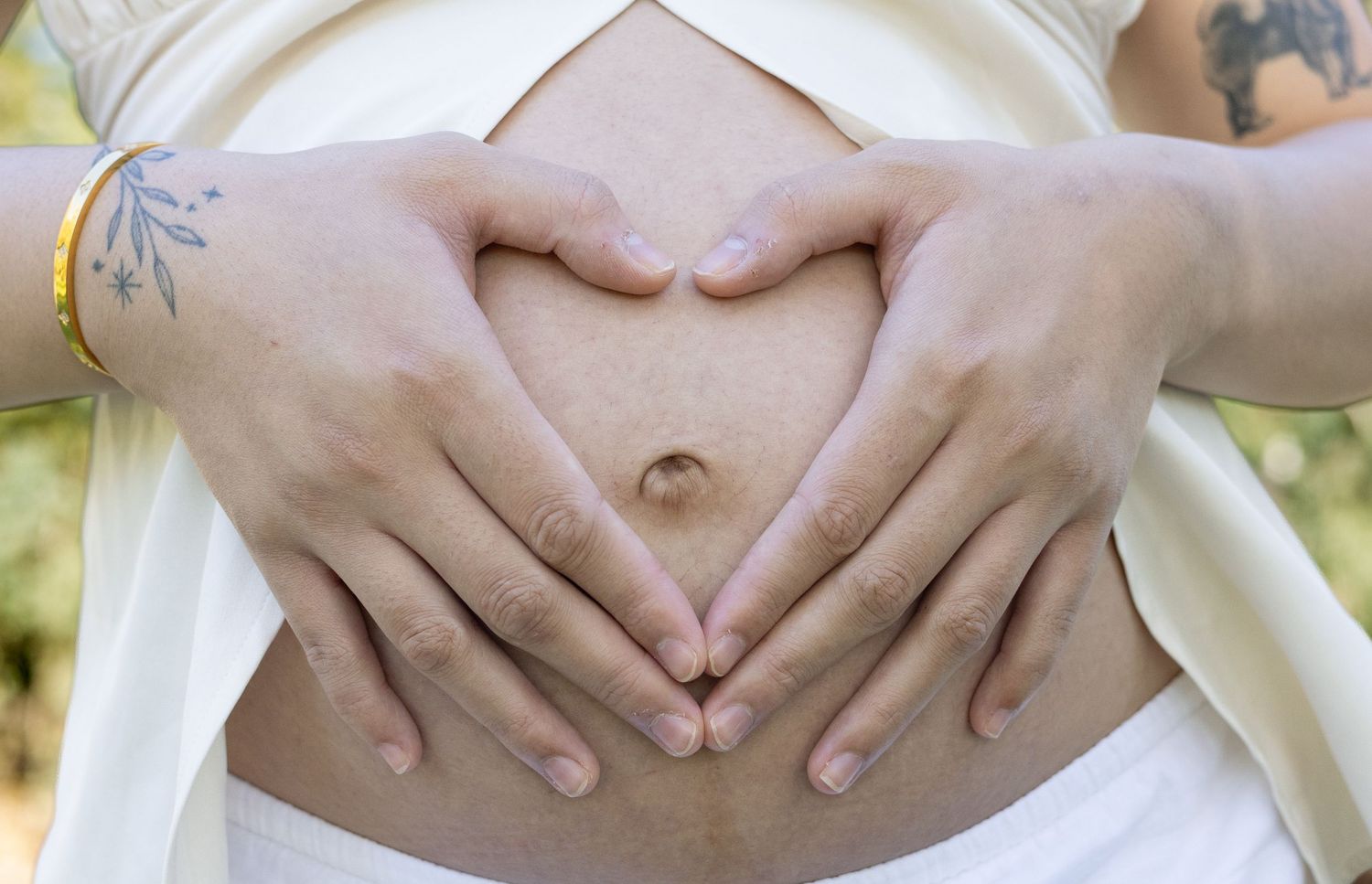 Pregnant woman in white strapless top and skirt forming heart shape with hands on baby bump, with tattoos and gold jewelry.