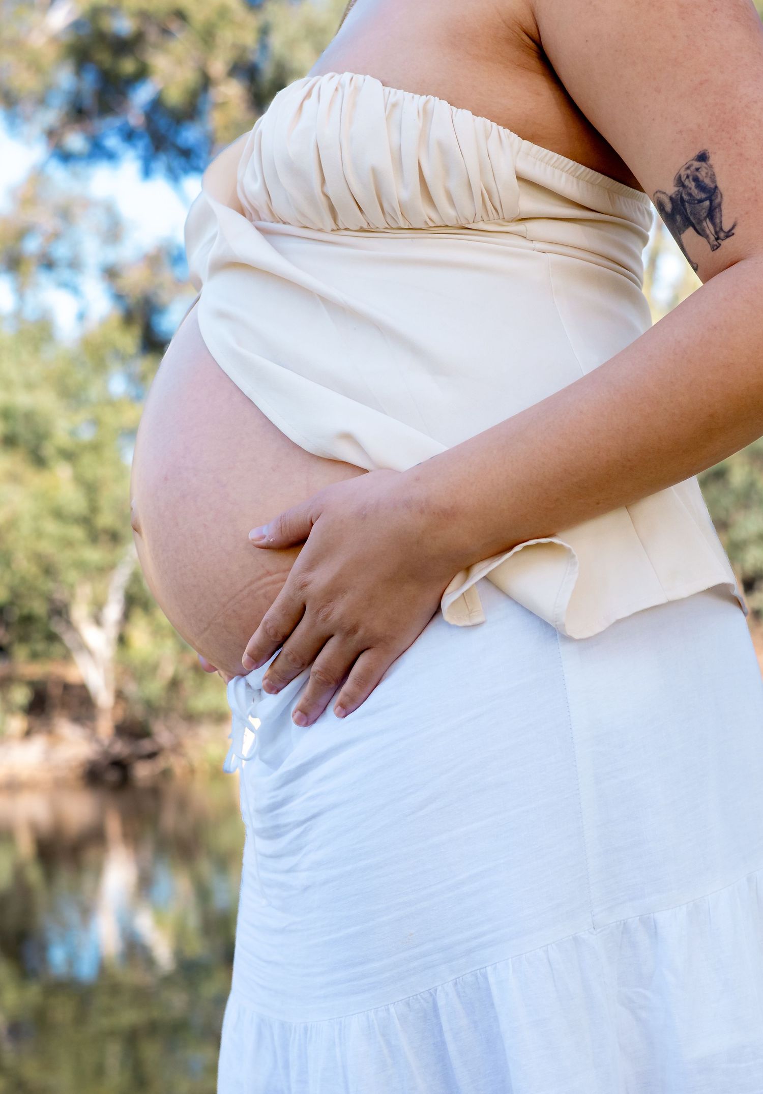 Pregnant woman in white strapless top and skirt forming heart shape with hands on baby bump, with tattoos and gold jewelry.