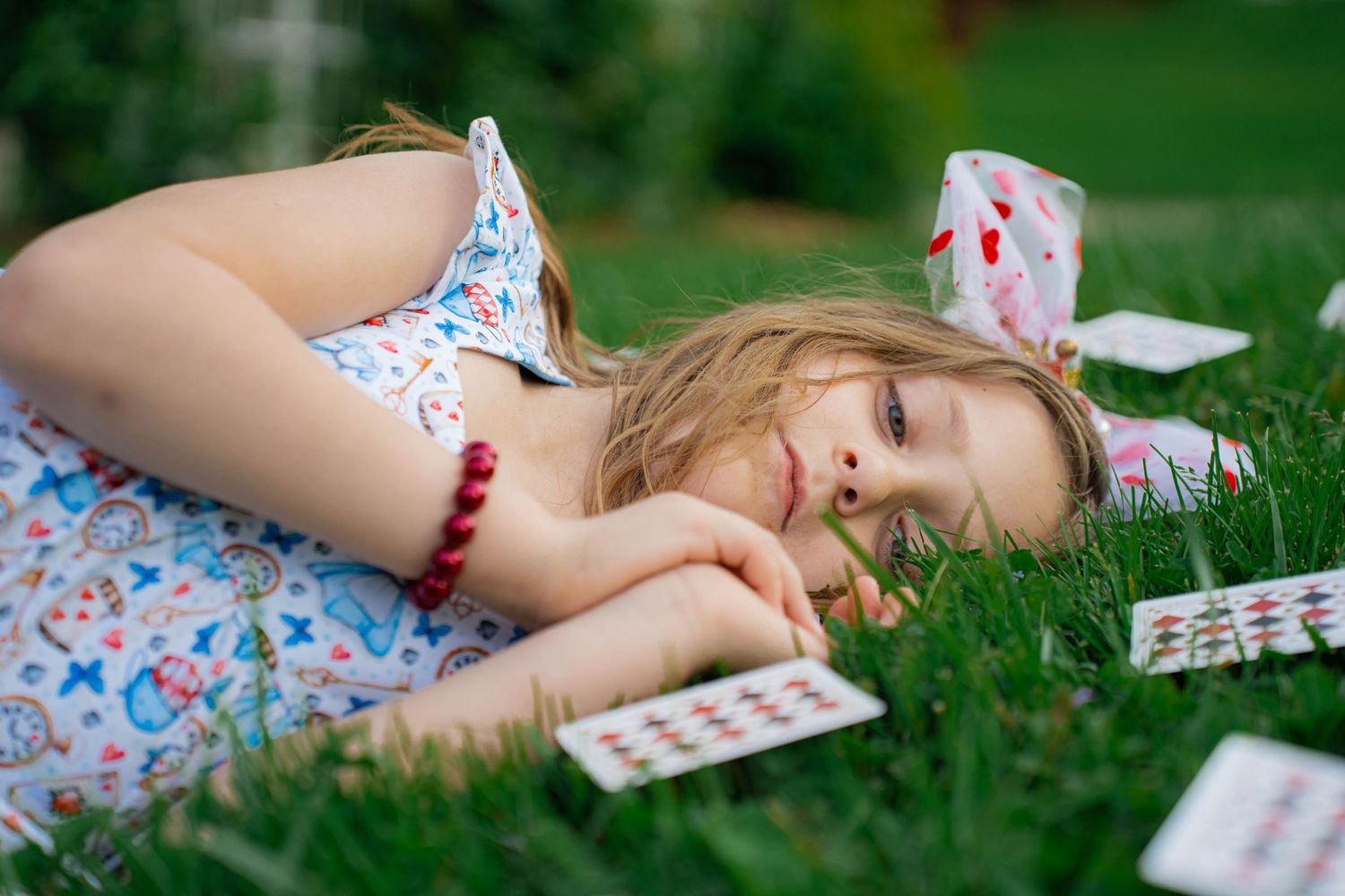 Young girl lying on green grass surrounded by scattered playing cards, wearing a floral dress and red bracelet.