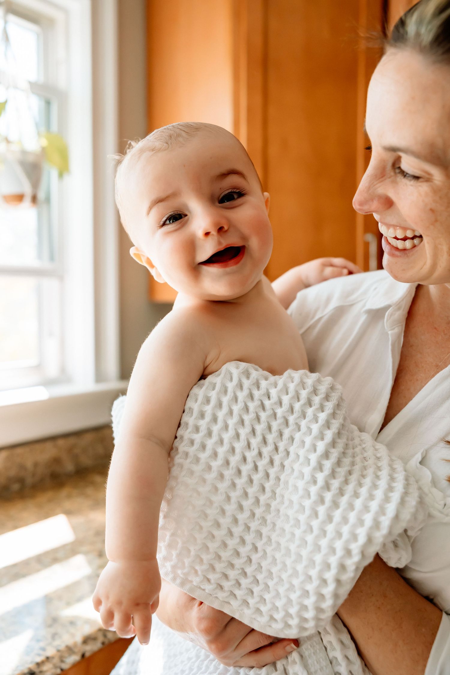 A happy baby wrapped in a white towel smiles while being held by a laughing woman in a bright kitchen.