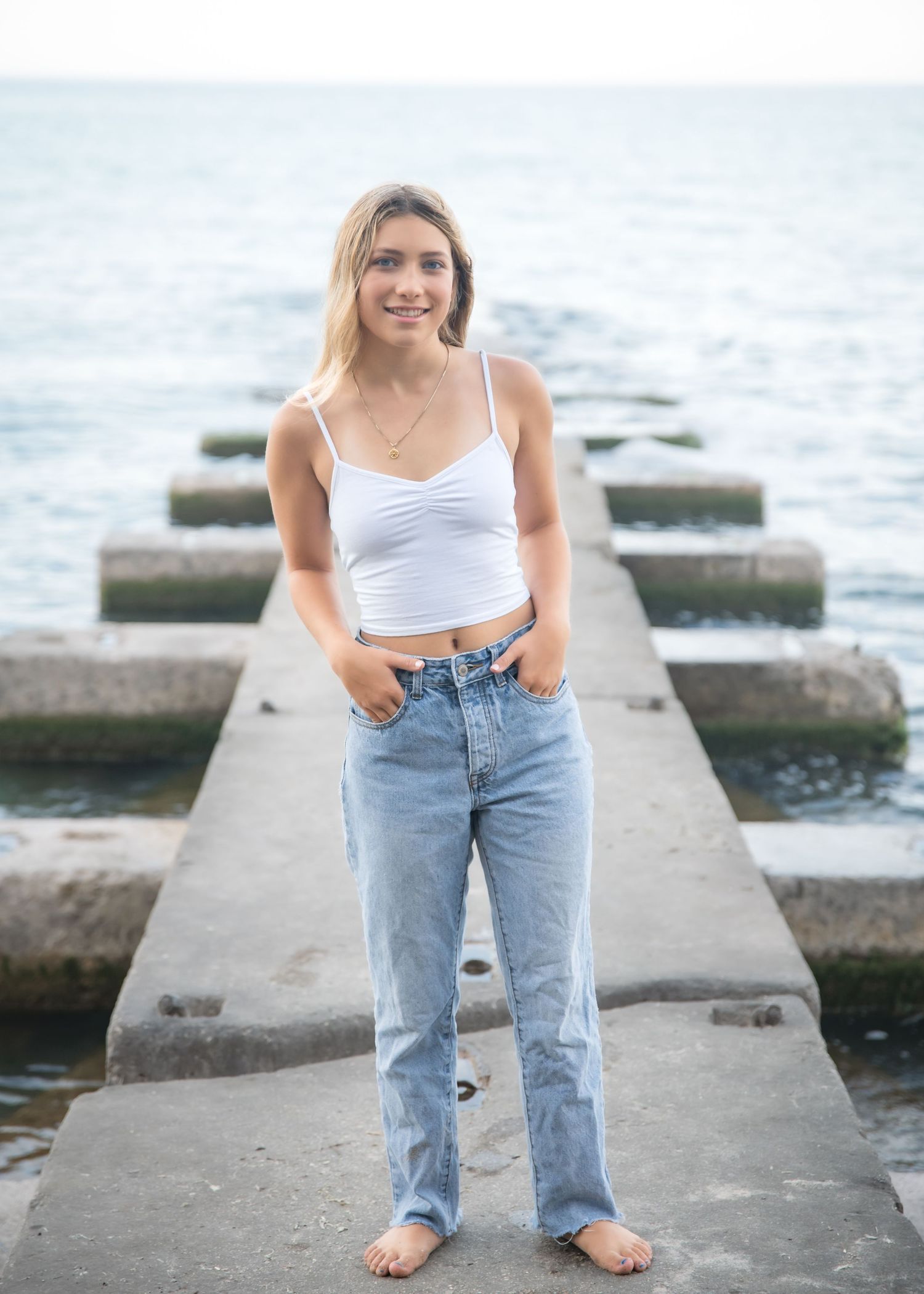 Young blonde woman in navy blue mini dress standing on driftwood log among large rocks on a beach.