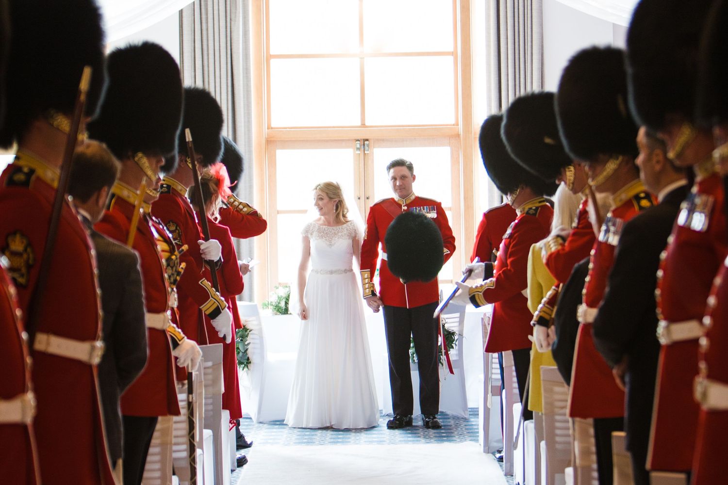 Military wedding ceremony with guards in red uniforms and bearskin hats lining the aisle as couple walks together.