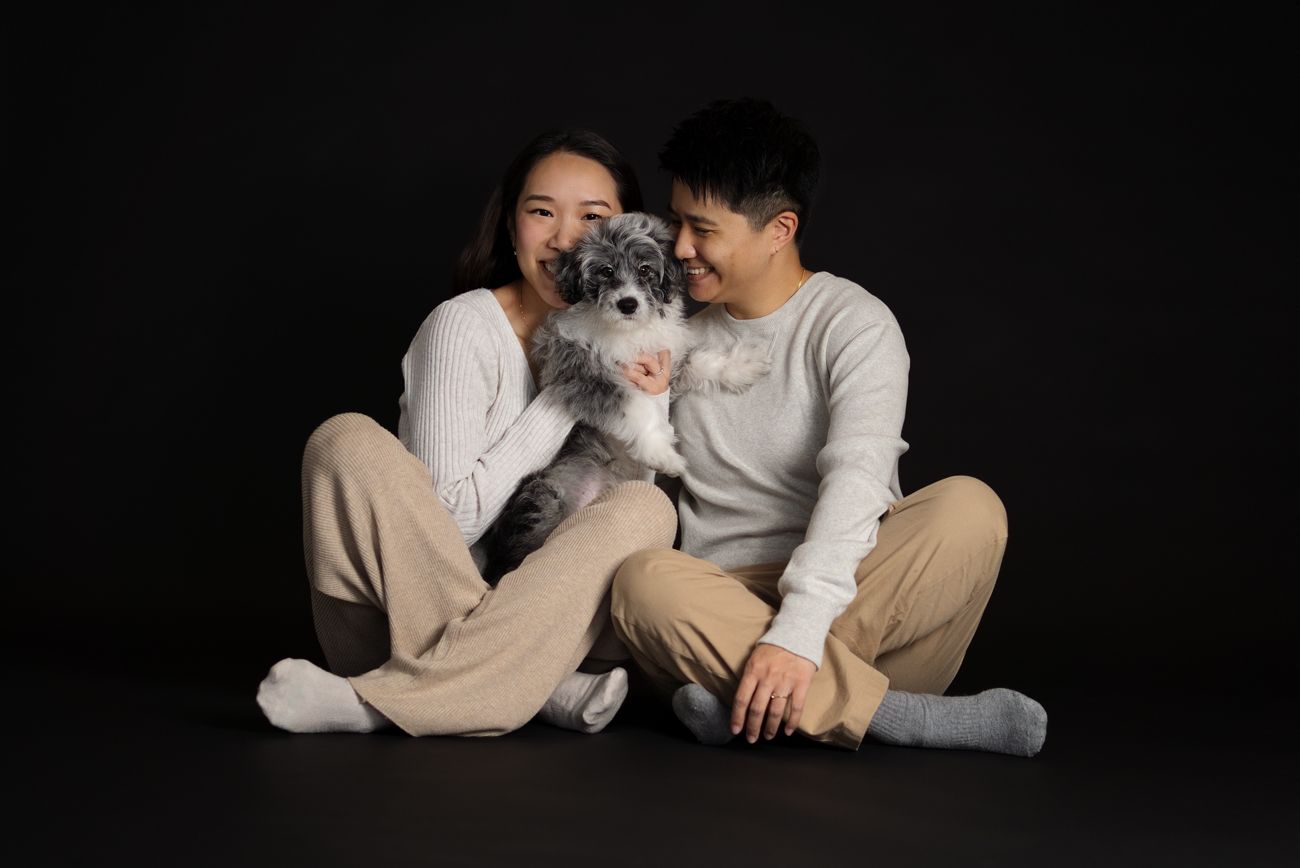 A couple in matching beige and white outfits sit on a dark floor, smiling while holding a fluffy gray and white dog in rouse hill sydney