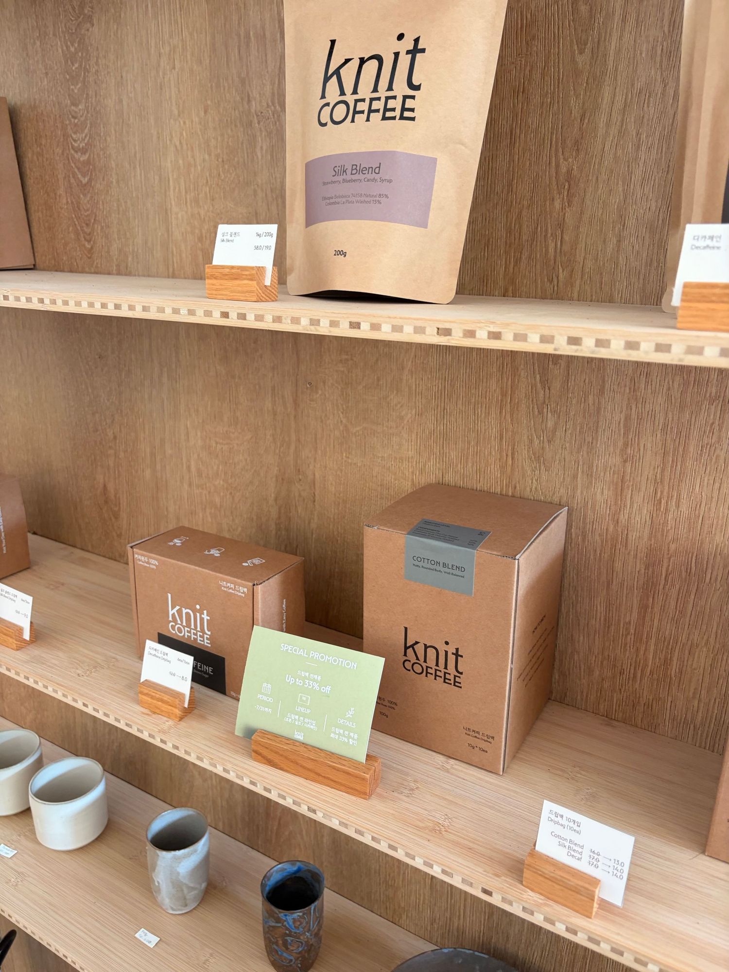 Knit Coffee branded bags and boxes displayed on wooden shelves alongside ceramic mugs in a minimalist cafe setting.