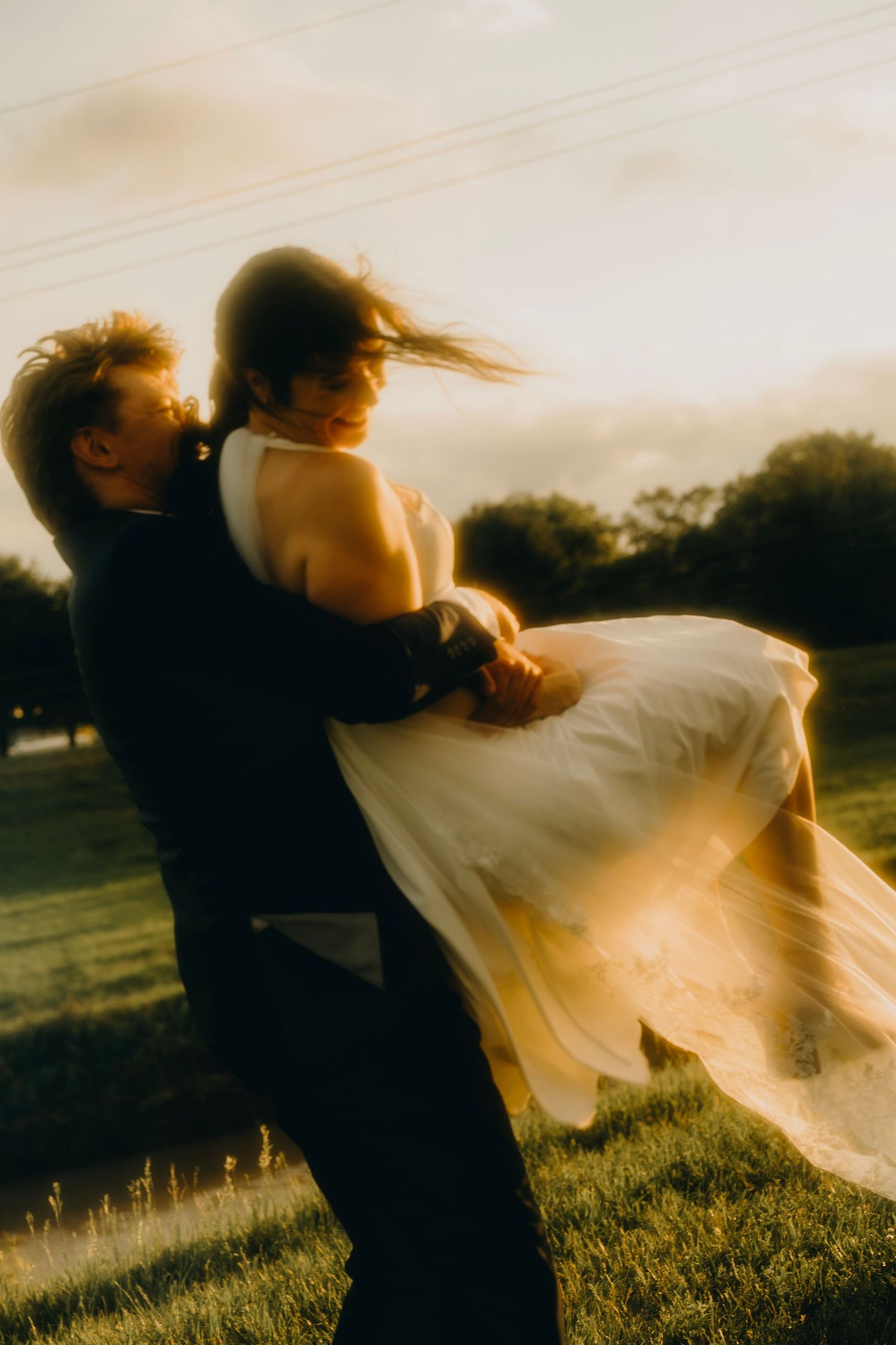 Groom lifting bride in golden sunset light, her white dress flowing in the wind during romantic outdoor wedding celebration.