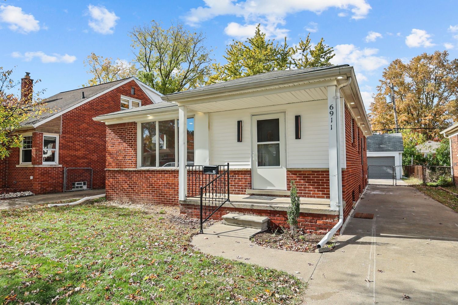Charming brick ranch home with white trim, covered front porch, and detached garage amid autumn trees.