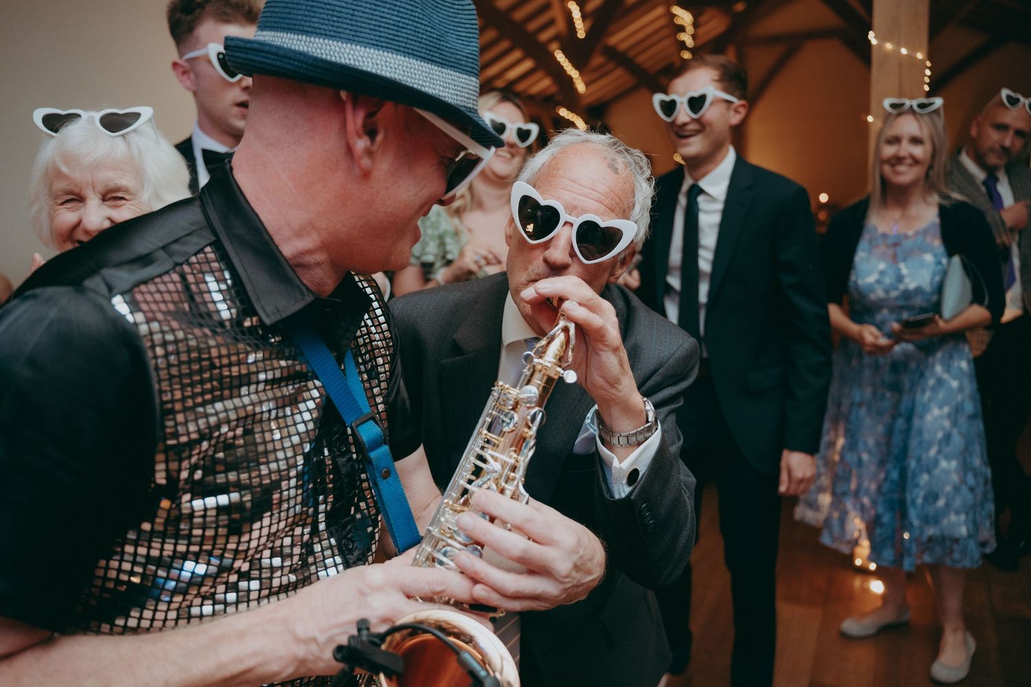 Man in sequin vest plays saxophone while guest in heart sunglasses leans in at lively wedding reception.