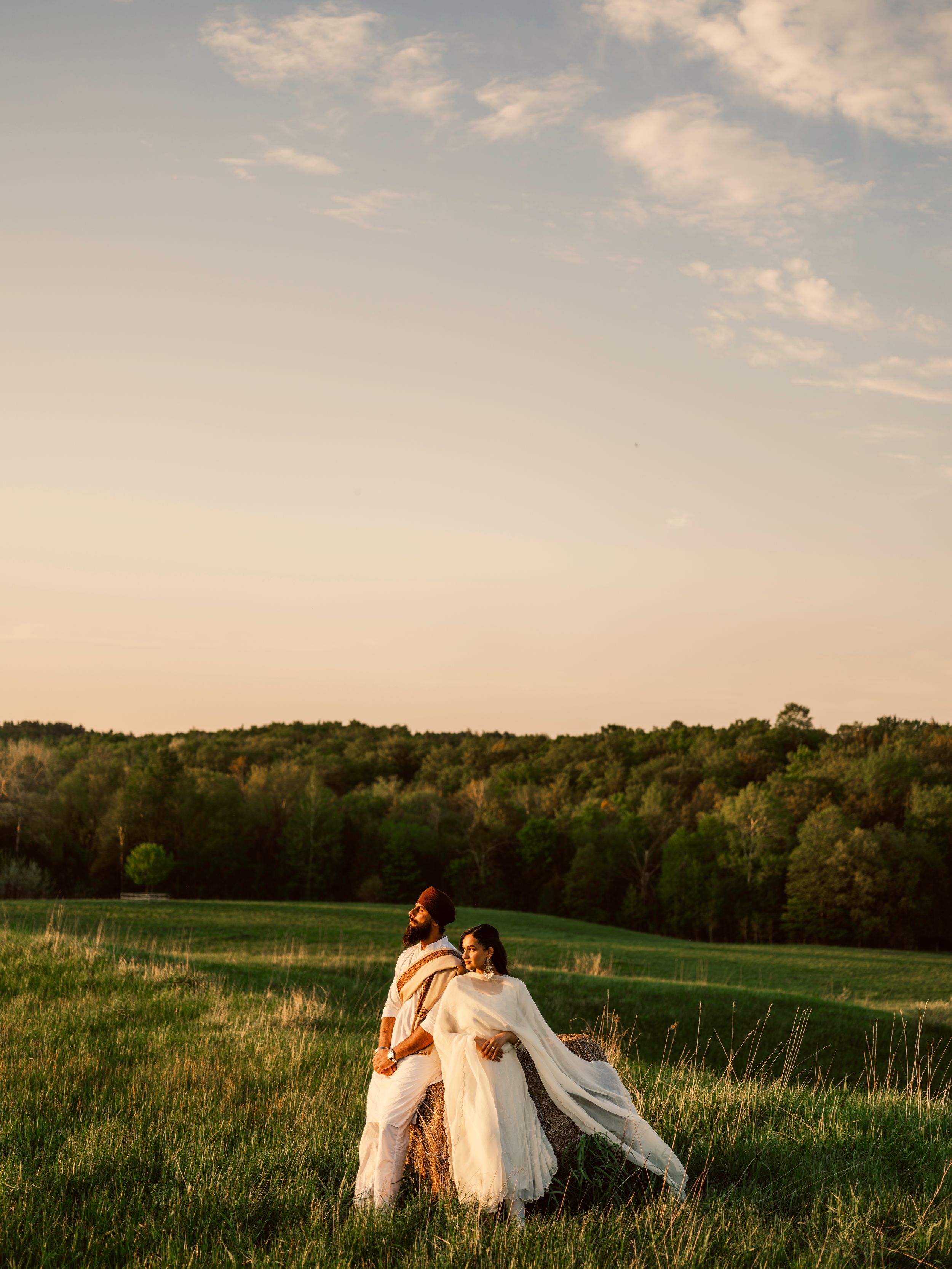 Couple in white attire embracing in a lush green field at golden hour with a forest backdrop.