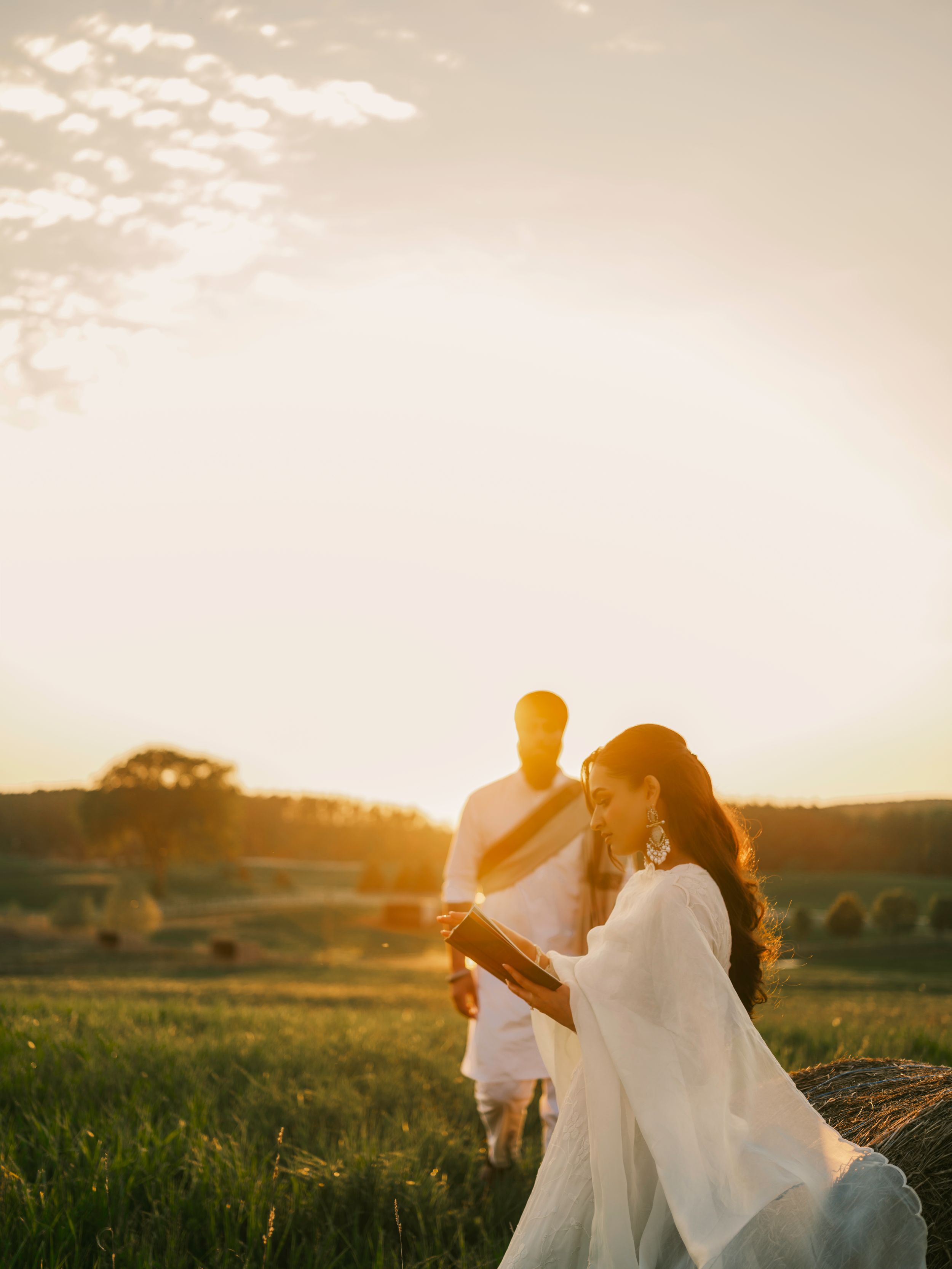 Couple in white attire during golden sunset in a lush green field with hay bales in warm evening light.