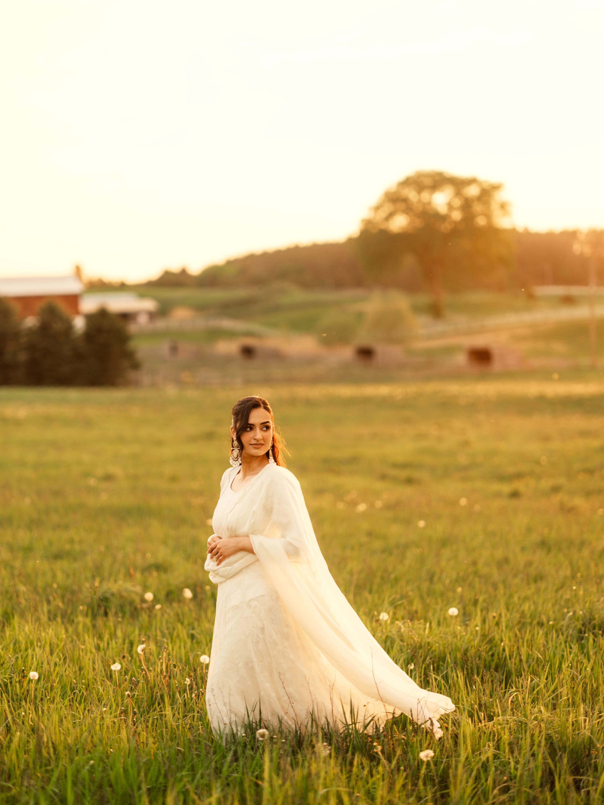 Bride in white gown standing in golden sunlit field at sunset, with rolling green hills and farm in background.