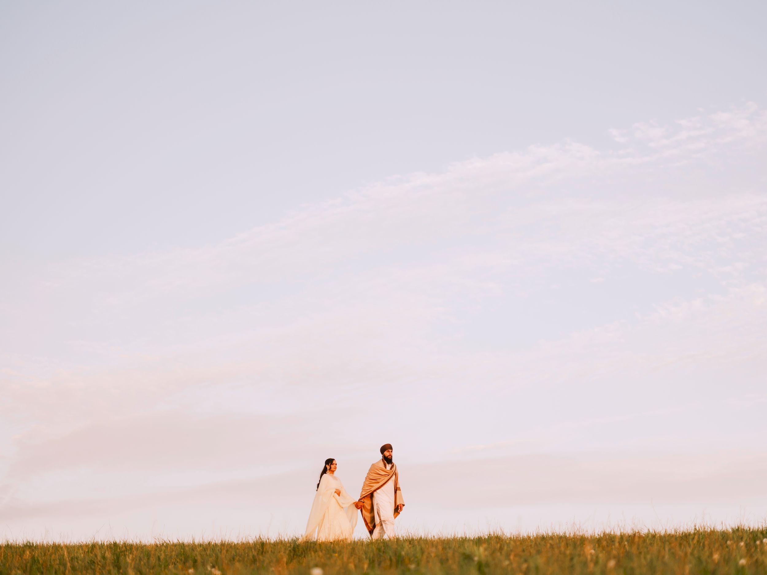 Couple in wedding attire walking hand in hand through a sunlit green meadow under a soft pastel sky.