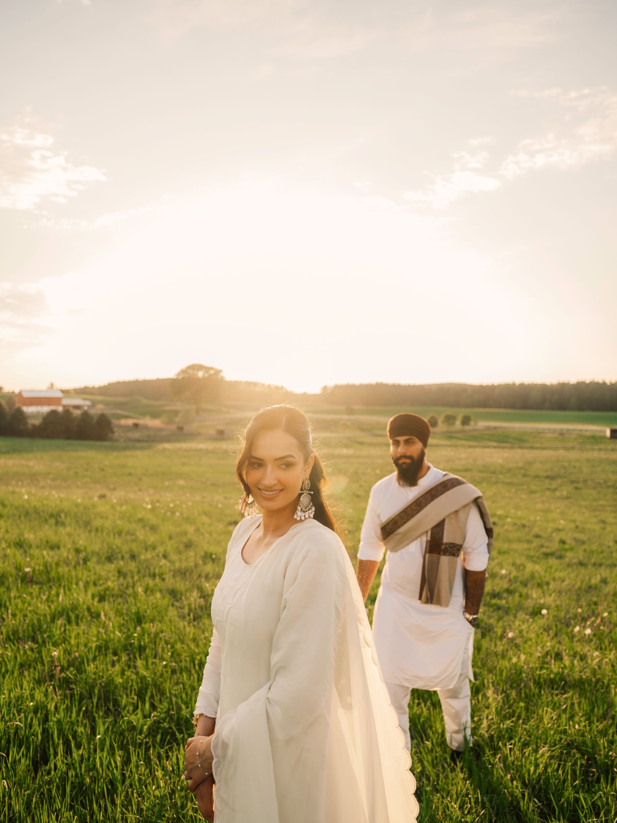 A couple in white traditional Sikh attire stands in a sunlit green field at golden hour.