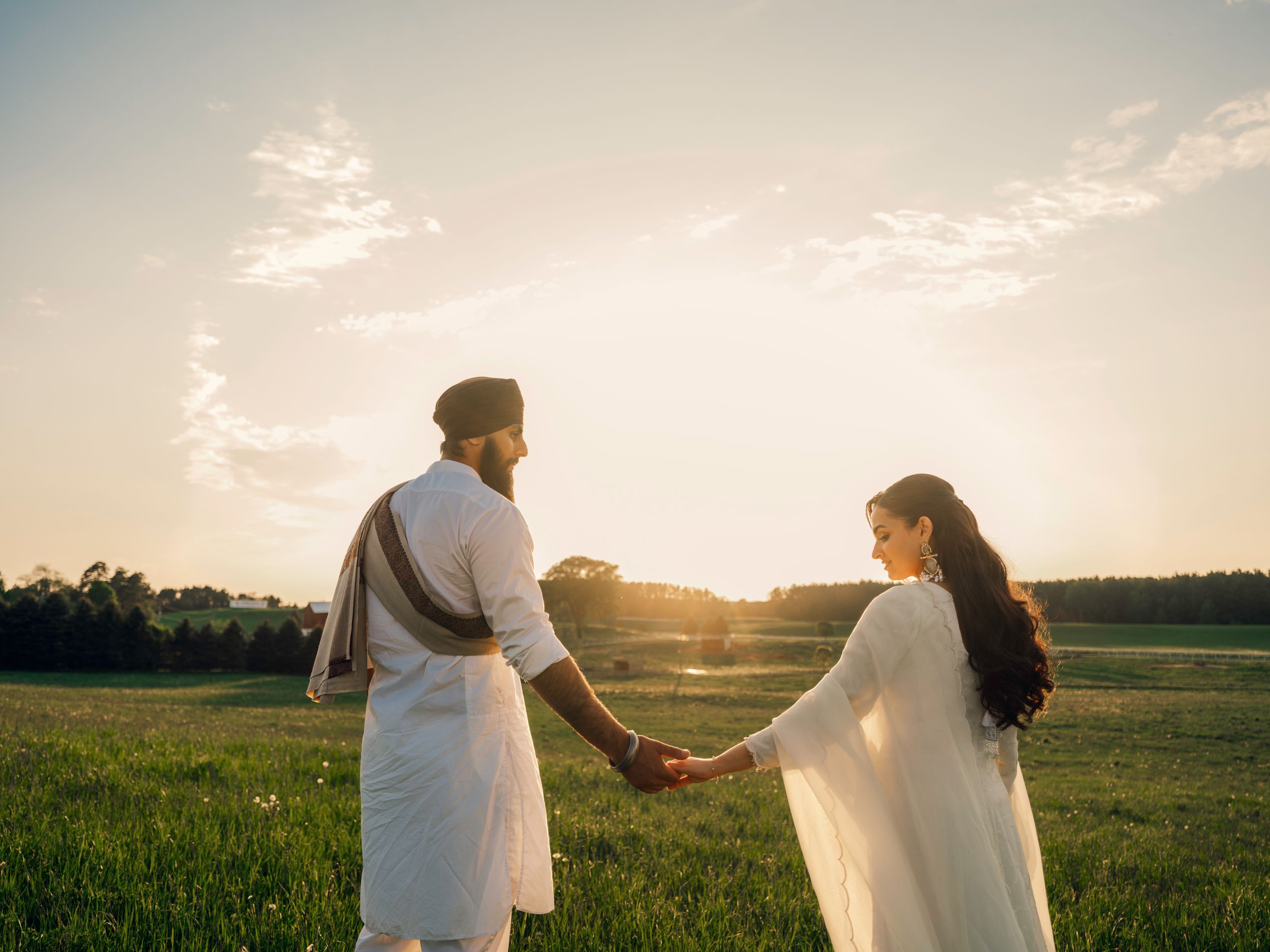 Sikh couple holding hands at sunset in a green field, wearing traditional white attire.