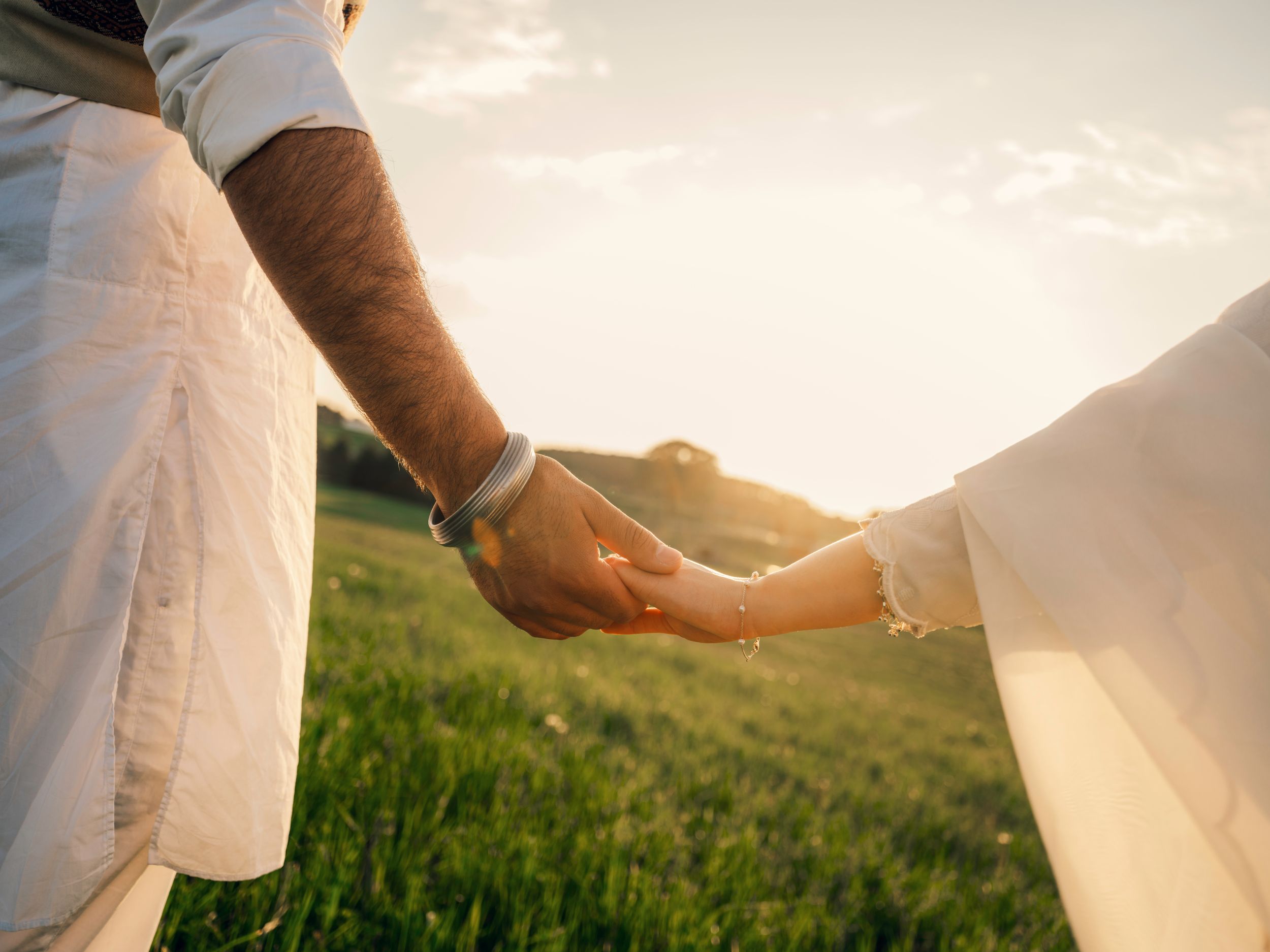 Couple holding hands in a green field at golden sunset, wearing white outfits, romantic outdoor moment.