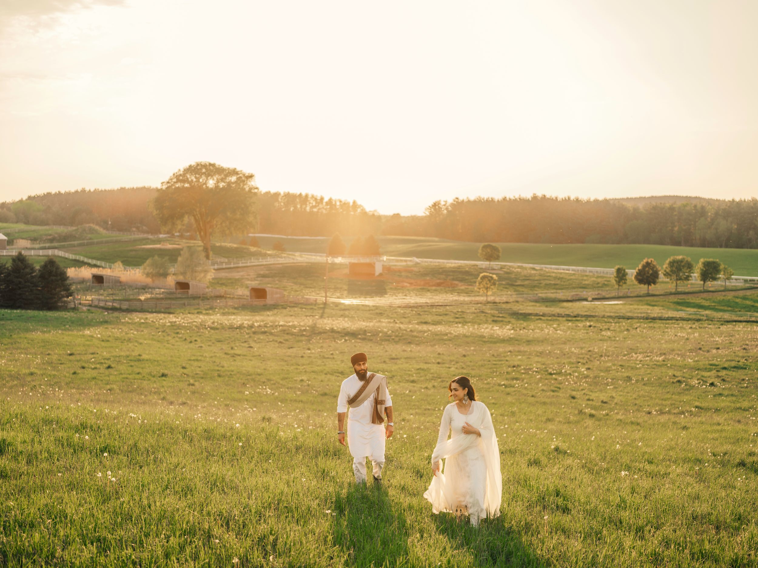 Couple in white wedding attire walking through golden sunlit green fields at sunset in a rural countryside landscape.