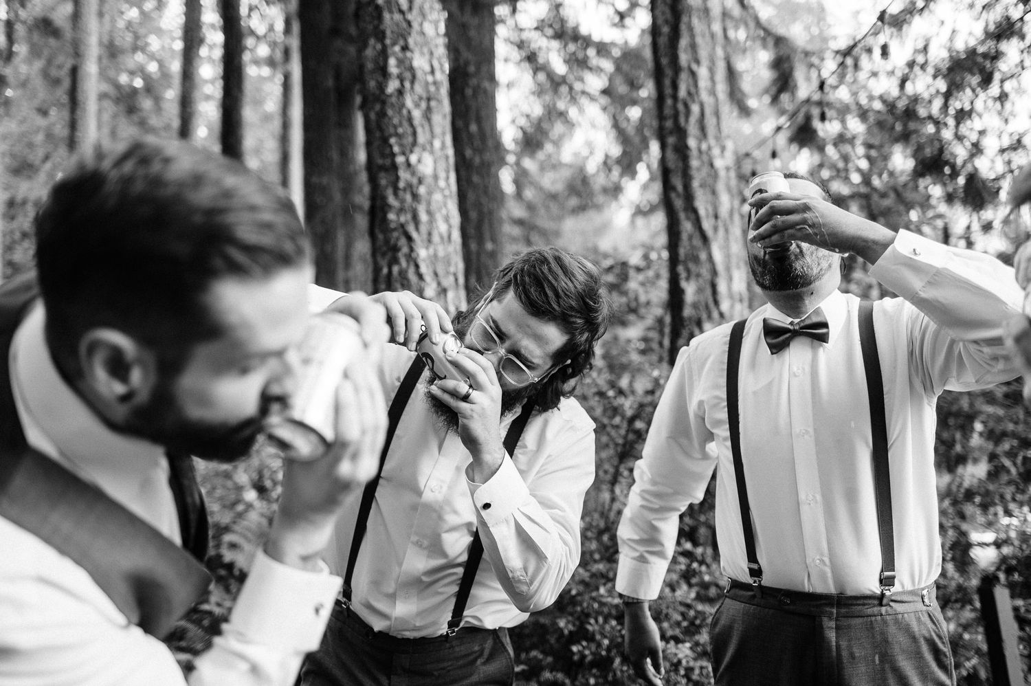 Black and white photo of groomsmen in suspenders and bow ties shotgunning beer in the outdoors among trees.