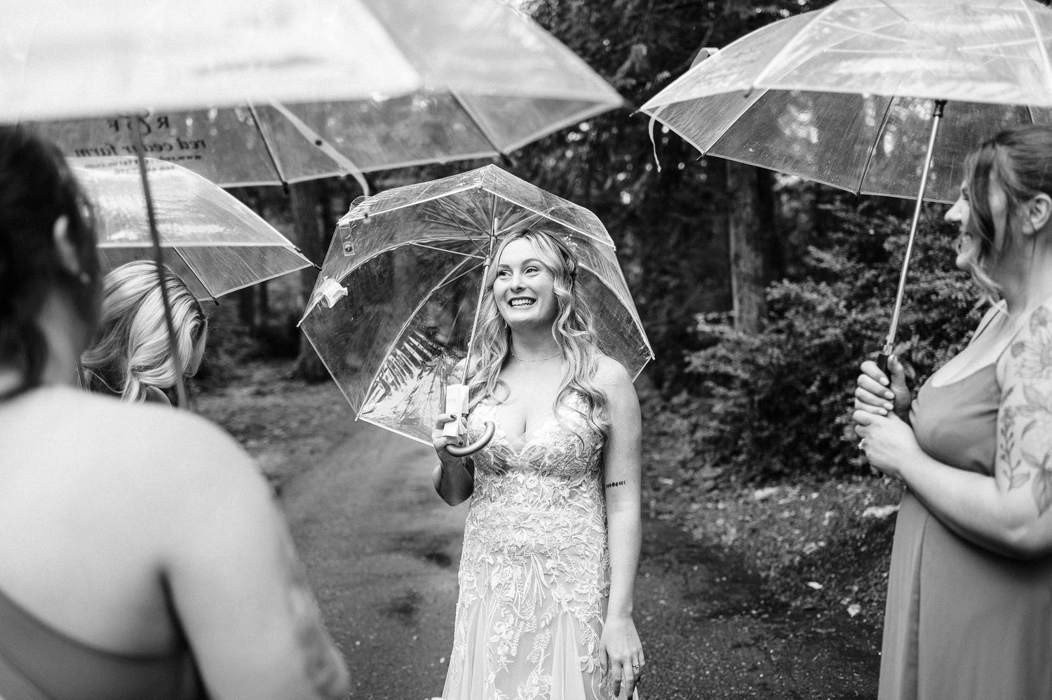 Bride laughing under clear umbrella with bridesmaids in rain, black and white wedding photo.
