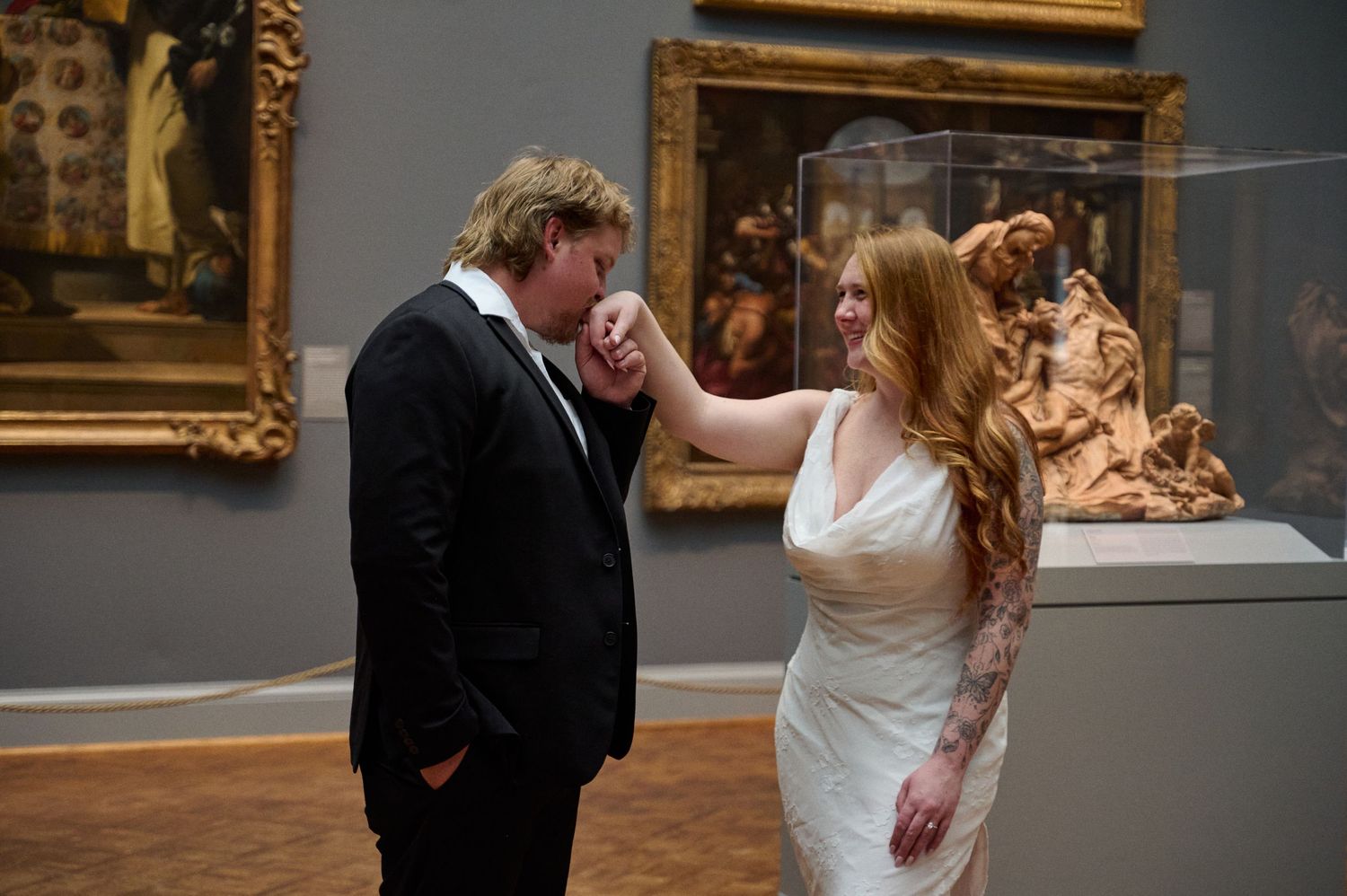 Groom kissing bride's hand in elegant art museum wedding portrait surrounded by classic paintings.