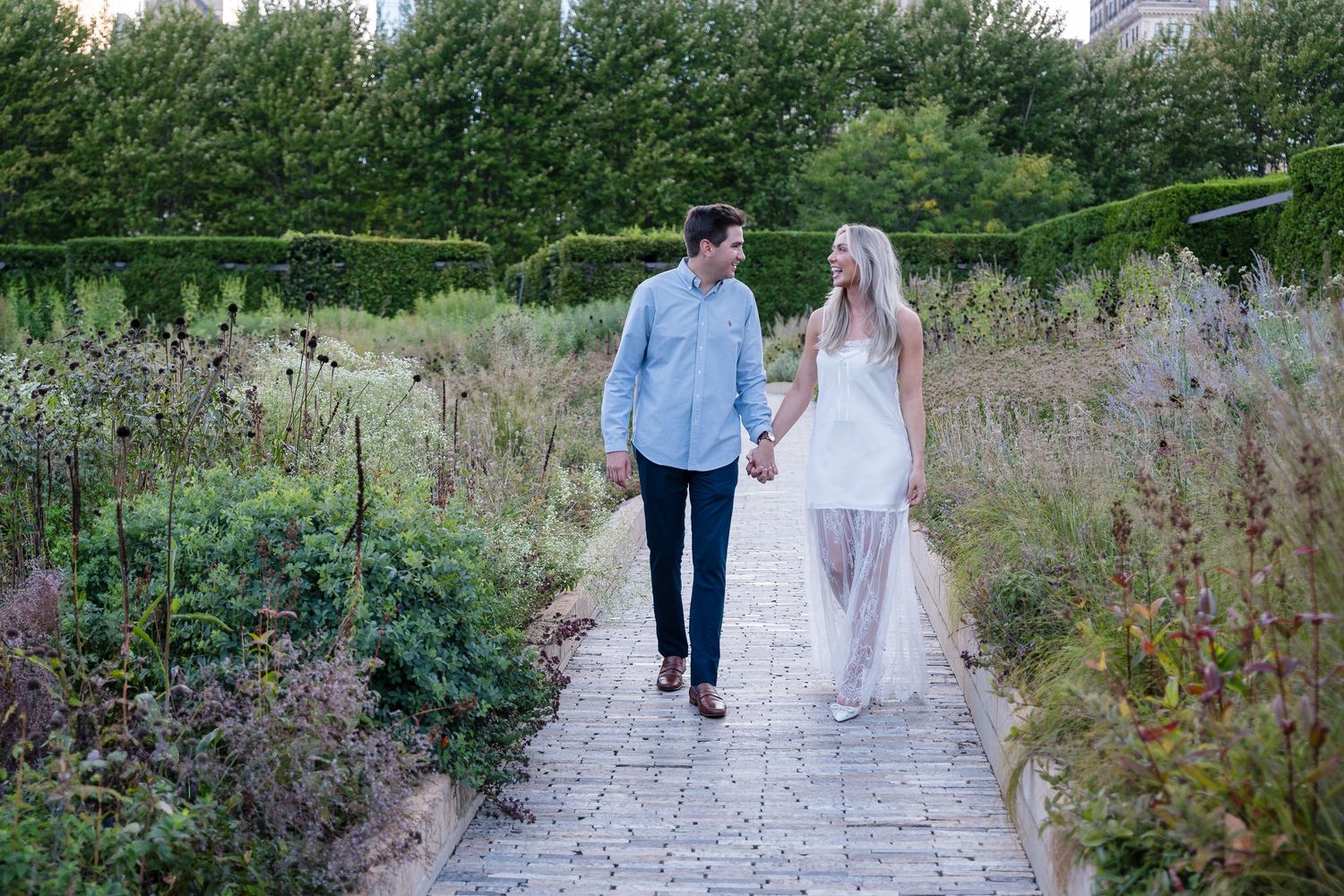 Couple holding hands walking on a brick garden path surrounded by lush greenery, wearing casual and white dress attire.