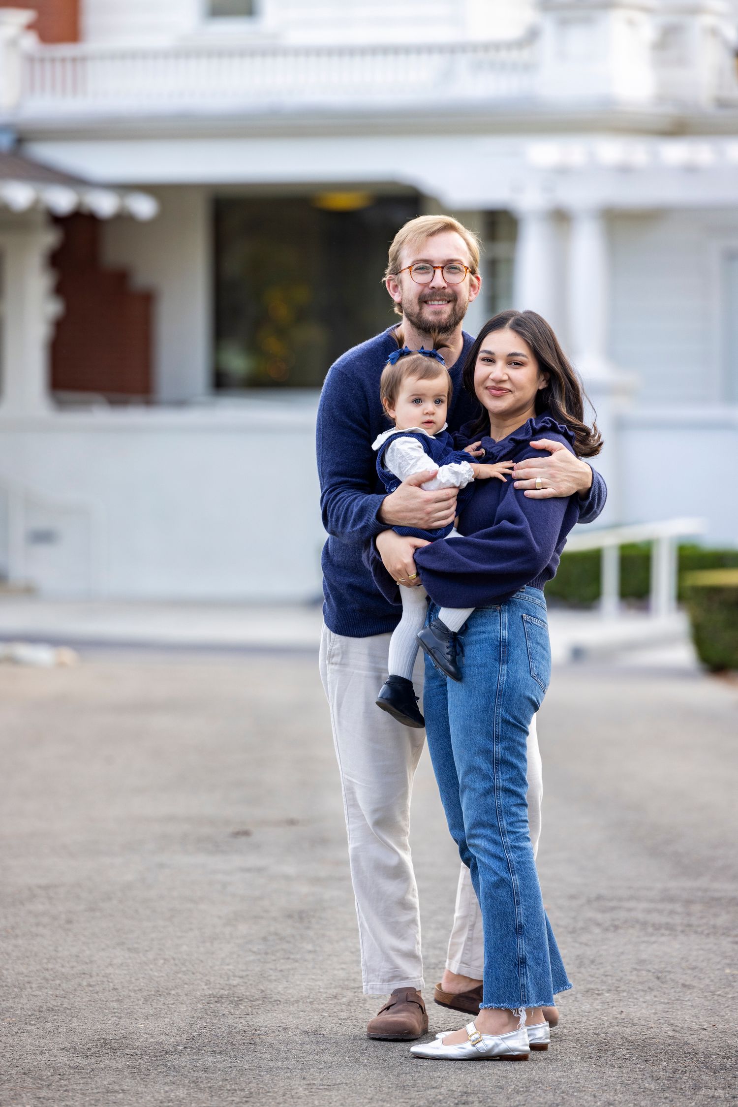 Happy family with baby posing in front of white house, man in navy sweater holding child with woman in jeans.