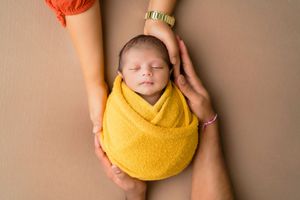 Newborn baby in yellow wrap held by parents' hands, studio photoshoot