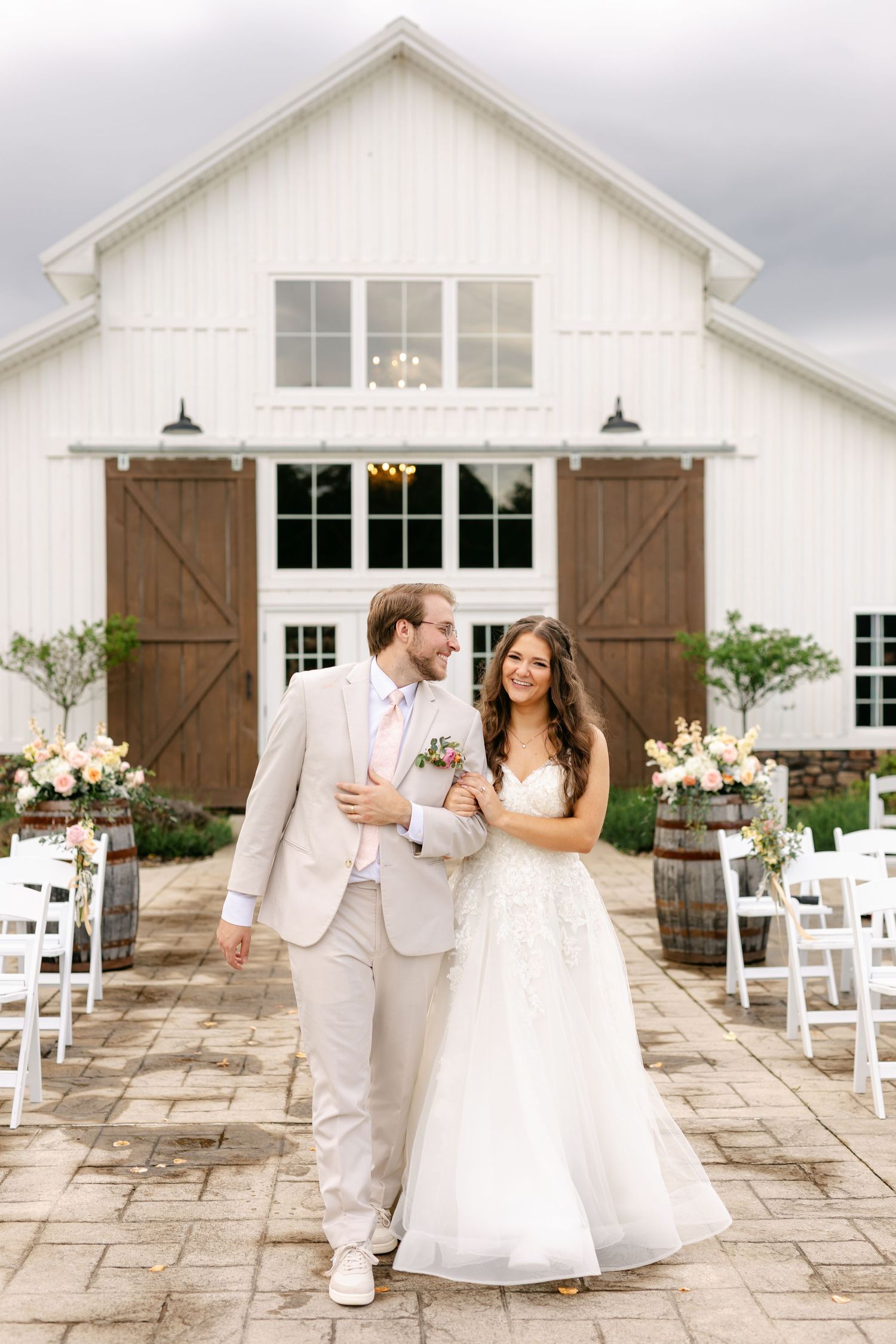 Happy bride and groom walking together in front of white barn venue with wine barrel floral arrangements.