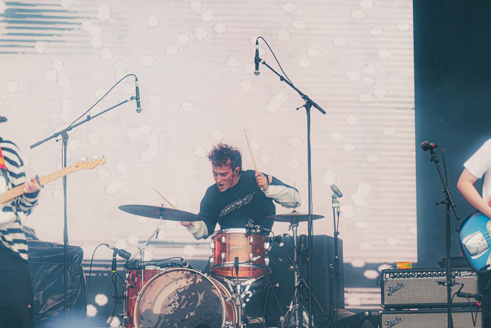 Drummer performing energetically on stage at a live music concert with band members and microphones visible.