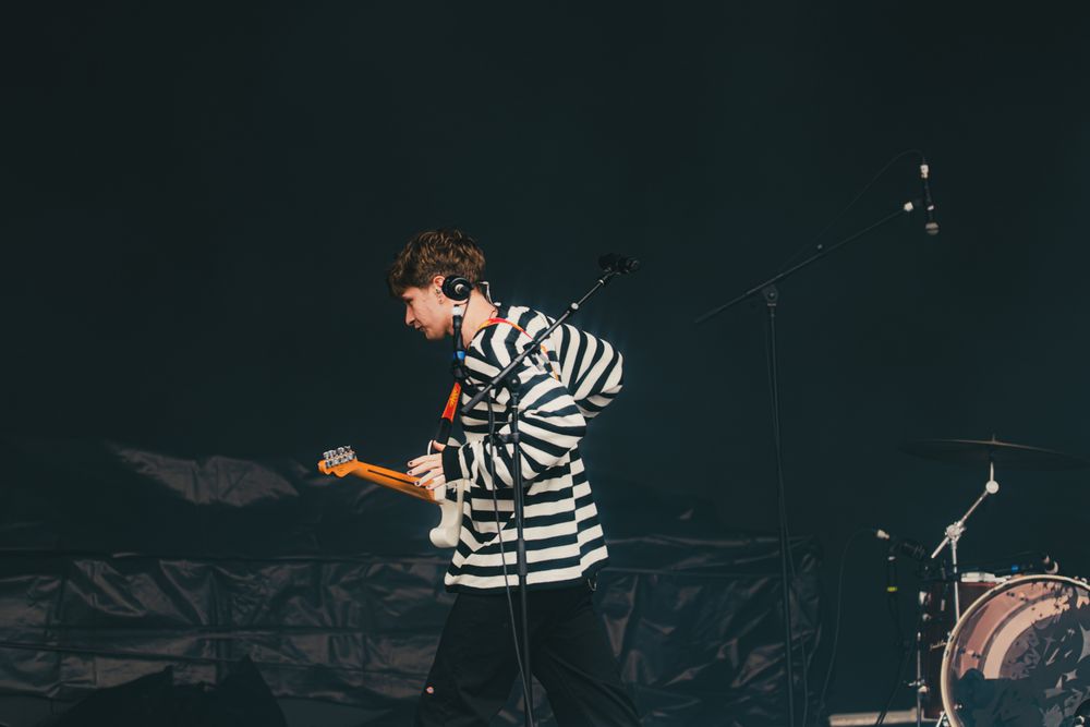 Guitarist in striped shirt performing on dark stage with electric guitar at live concert.