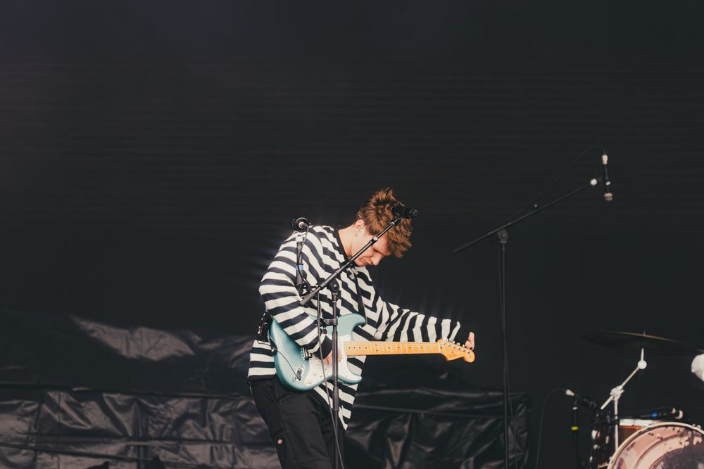Guitarist in striped shirt performing on dark stage with microphone stand during live concert.
