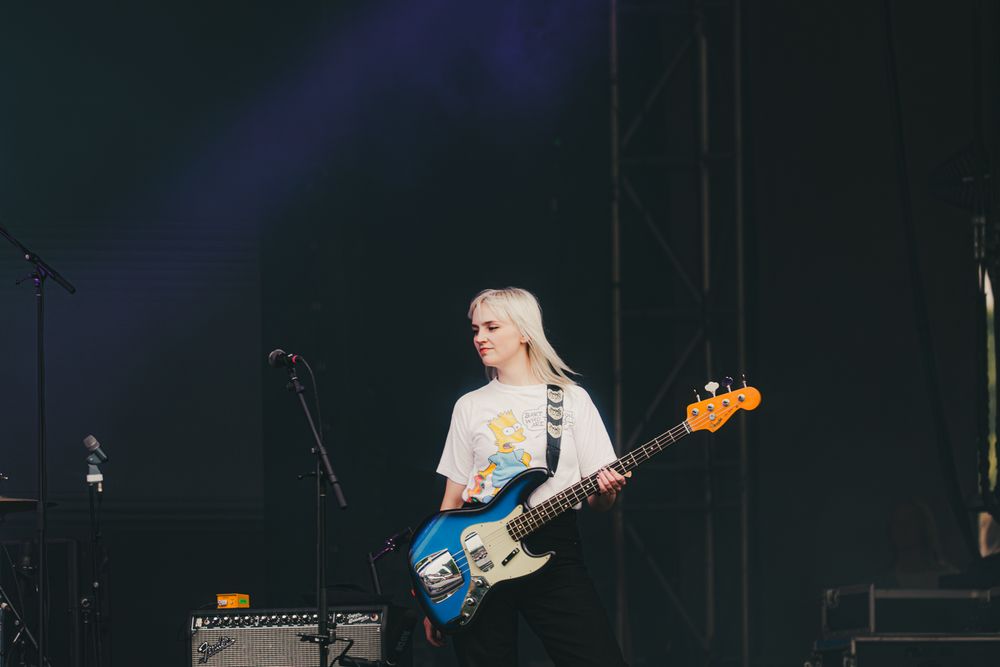 Blonde female guitarist performing on stage with a blue electric guitar at an outdoor music festival.
