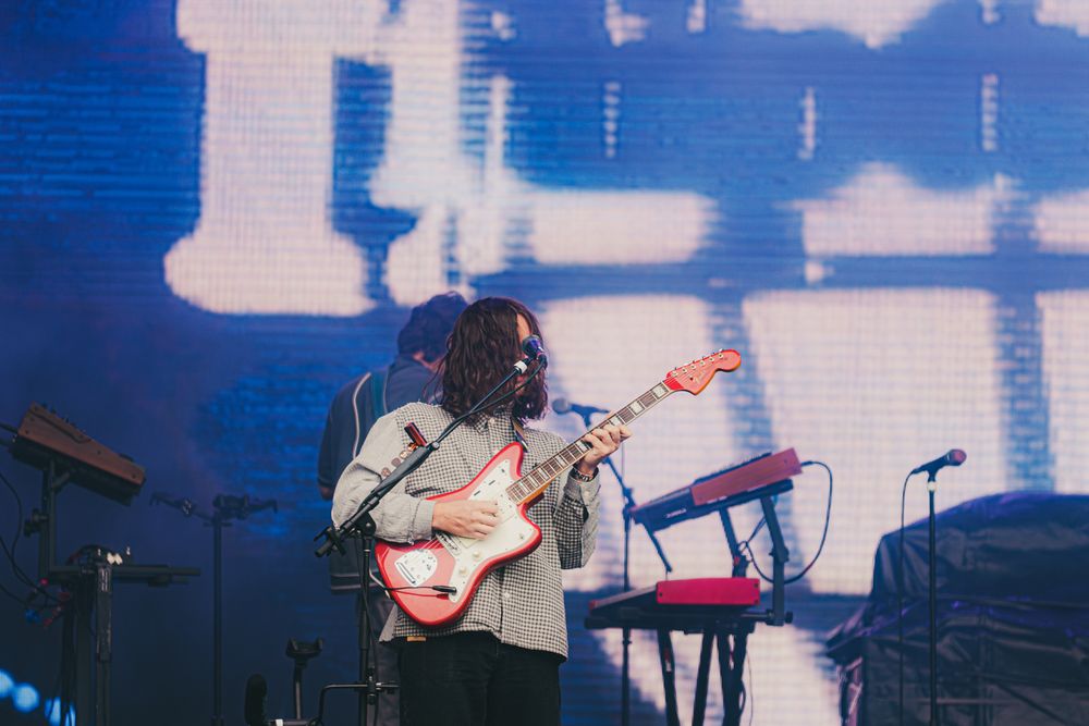 Guitarist performing on stage with a red electric guitar in front of a large illuminated screen at a live concert.