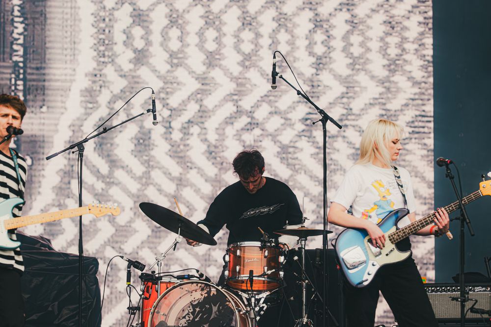 Band performing live on stage with guitarist, drummer, and blonde female guitarist in front of patterned backdrop.