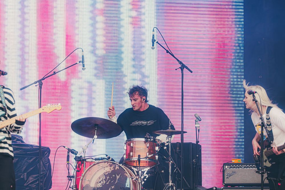 Band performing on stage with drummer in center, guitarist on left, and colorful LED backdrop at live concert.