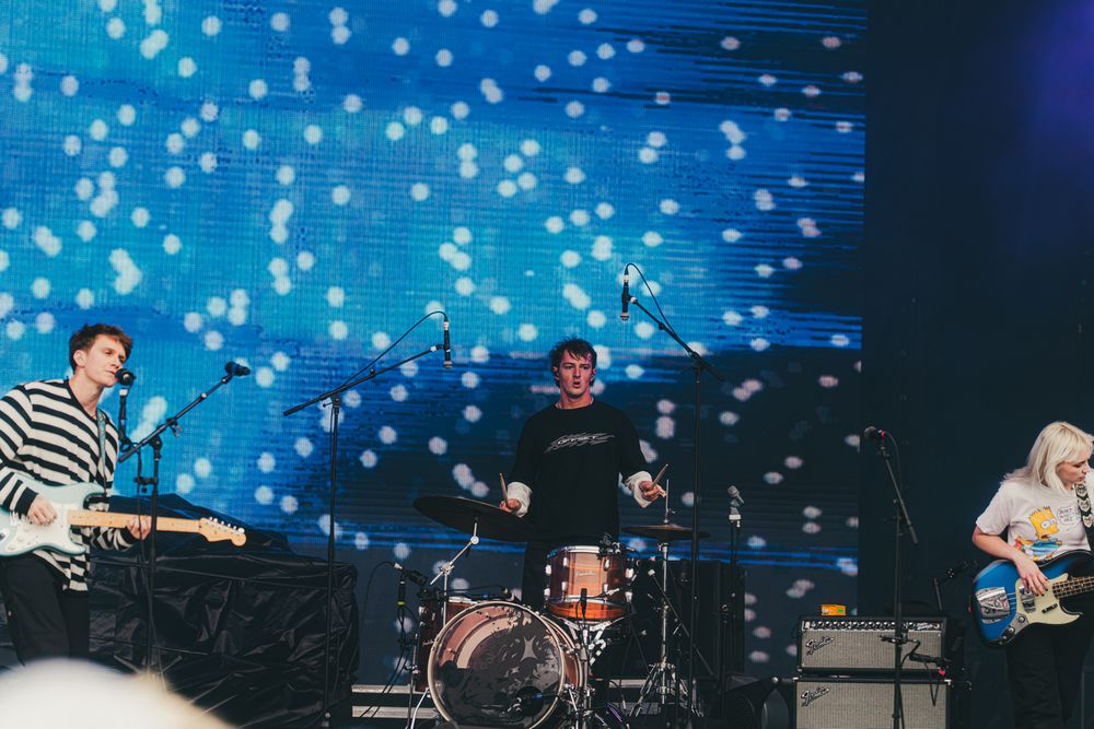 Band performing on stage with bokeh light backdrop, featuring guitarist, drummer, and bassist under blue spotlights.