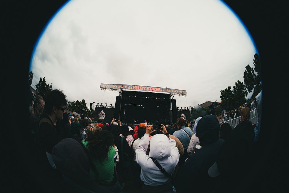 Fisheye lens view of a large outdoor music festival crowd facing a lit stage under an overcast sky.