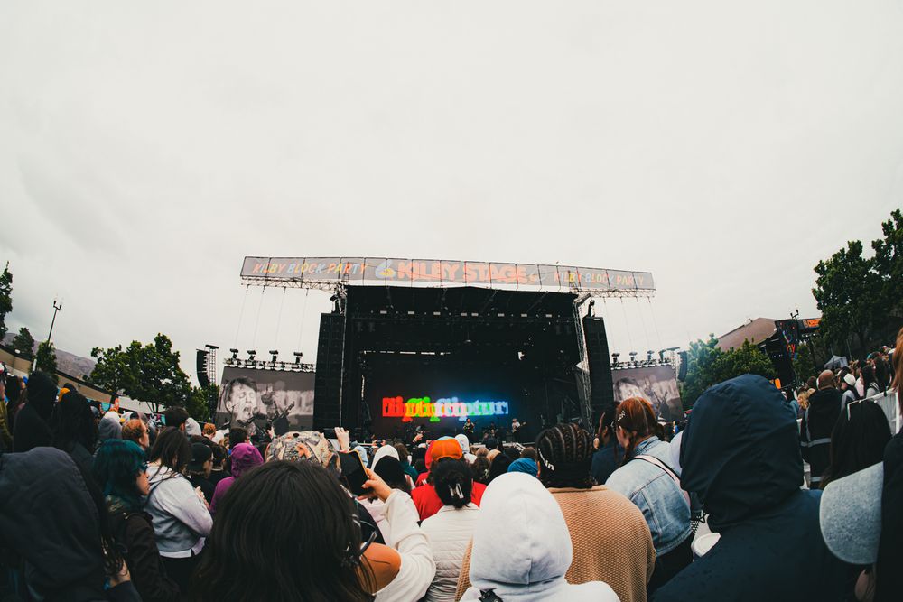 Crowd gathered at an outdoor music festival stage with colorful LED display on overcast day.