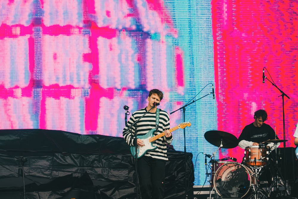 Guitarist performs on stage with colorful pink LED backdrop while drummer plays behind him at outdoor concert.