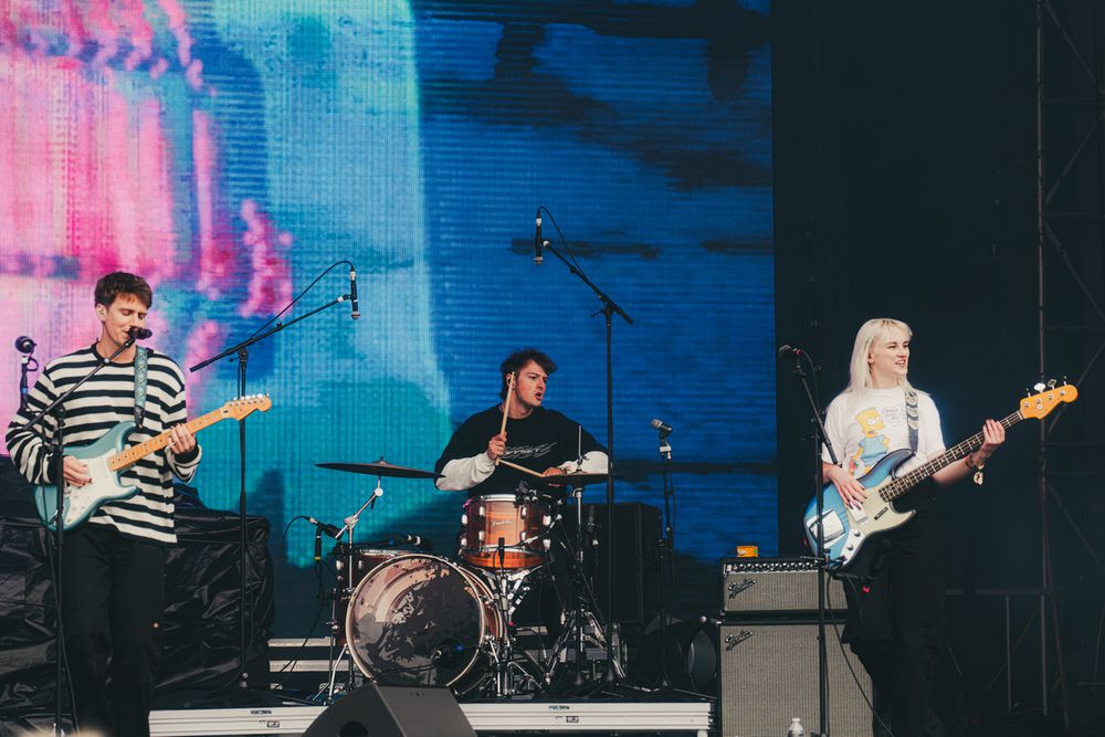 Three-piece band performs on outdoor festival stage, featuring two guitarists and a drummer with a blue projection screen behind them.
