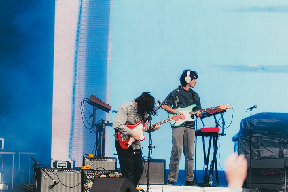 Two guitarists perform energetically on a festival stage, one in headphones at keyboard, with a blue-lit backdrop and crowd visible.
