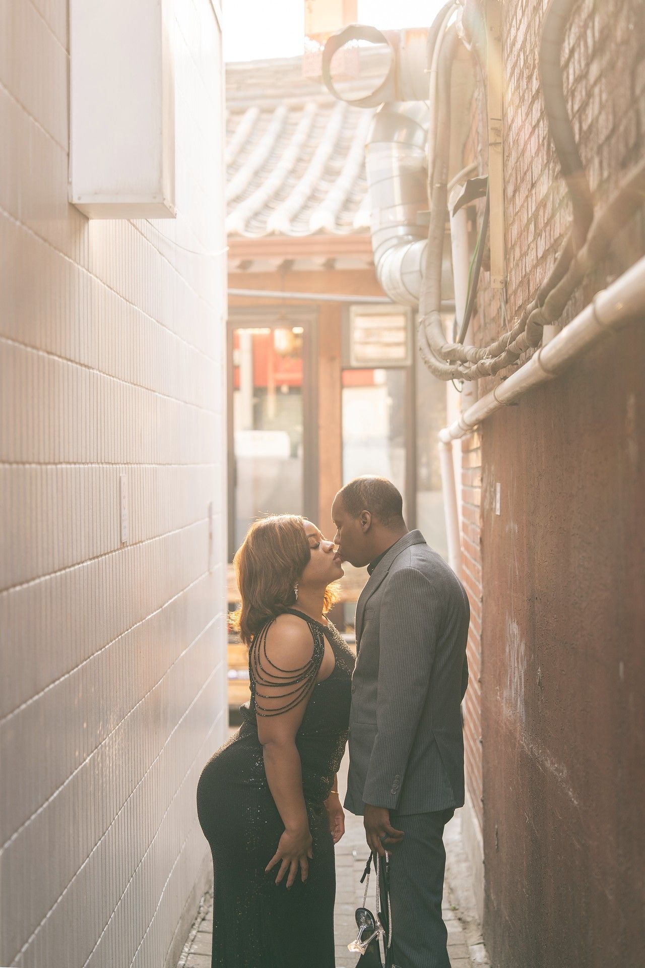 Couple sharing a romantic moment in a sunlit urban alley, woman in black dress holding a rose.