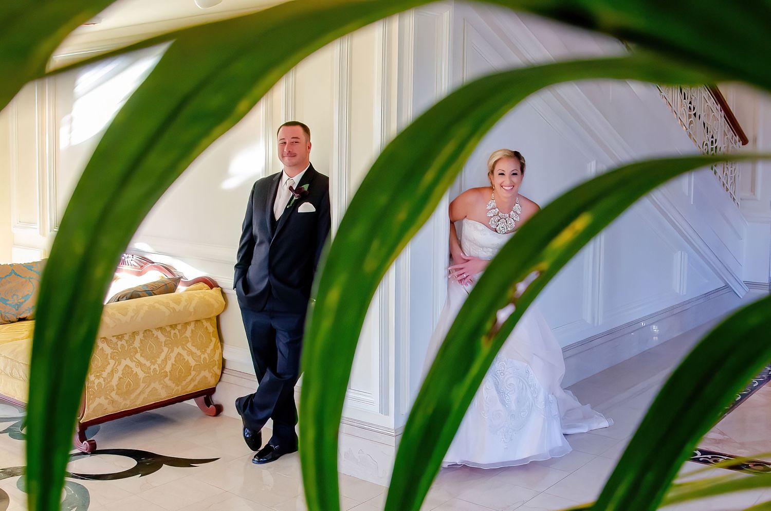 A bride and groom stand apart in an elegant venue, framed by lush green tropical leaves in the foreground.