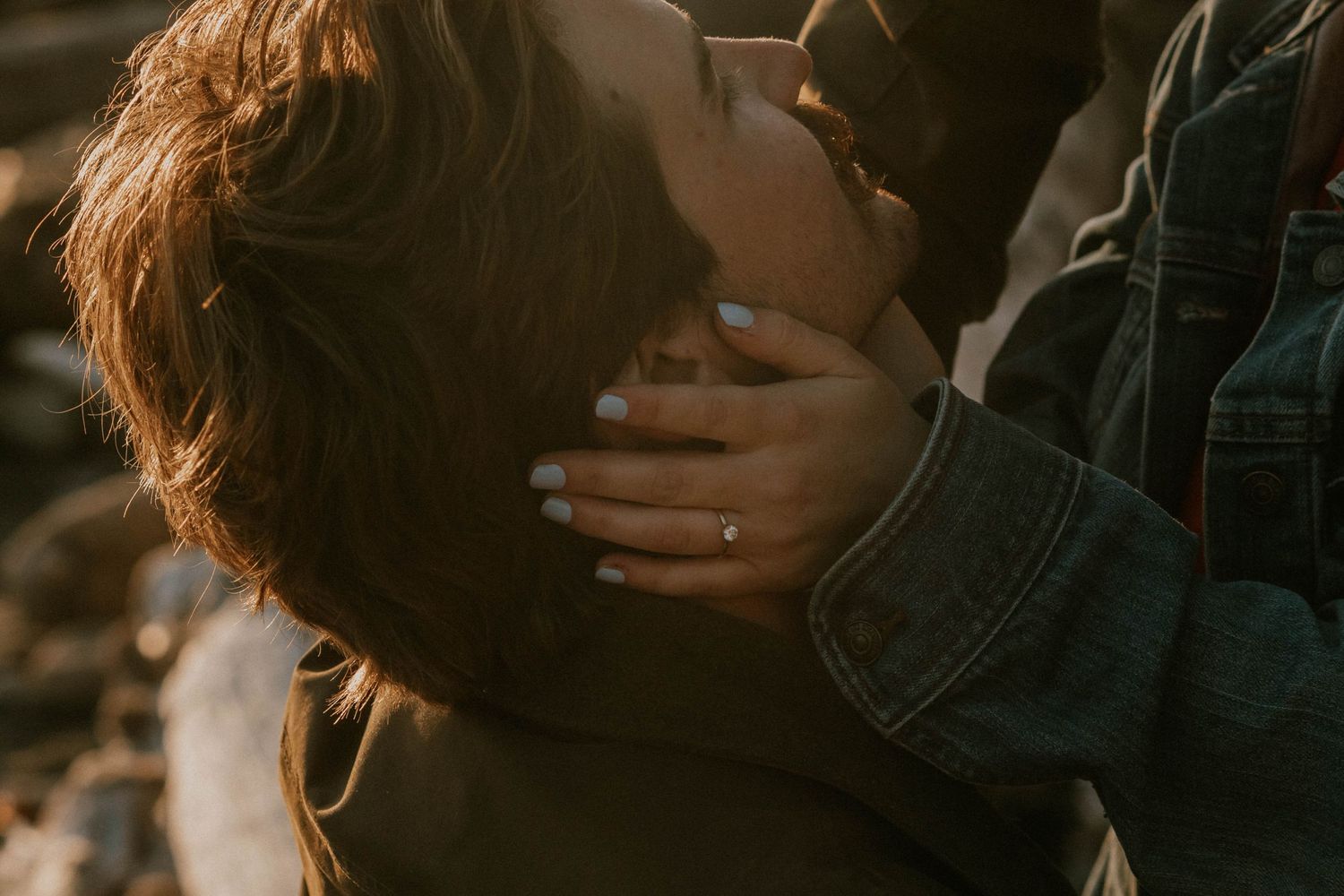 Close-up of a couple sharing a tender embrace, woman wearing an engagement ring, bathed in warm golden hour light.