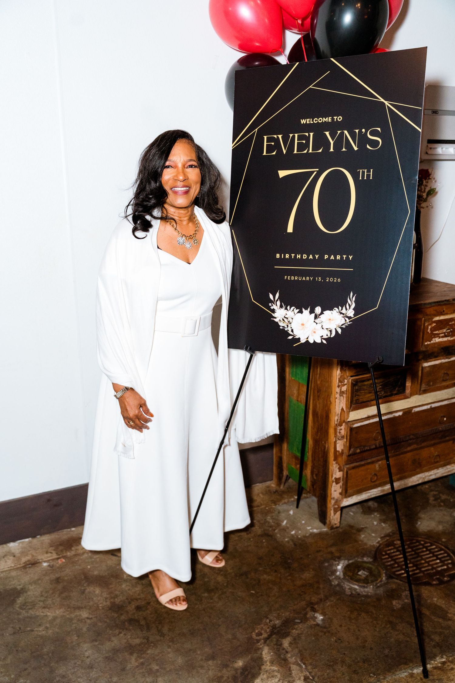 Woman in white dress poses beside Evelyn's 70th Birthday Party welcome sign with red and black balloons.