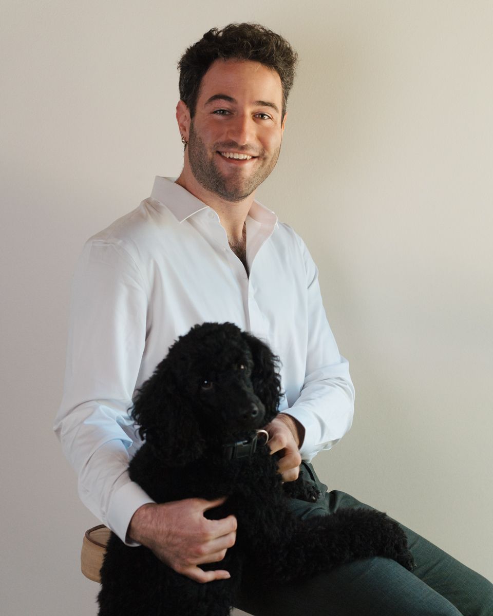 Man in white shirt sitting on stool, smiling while holding a black poodle dog against light background. Dog photoshoot in the Hills district