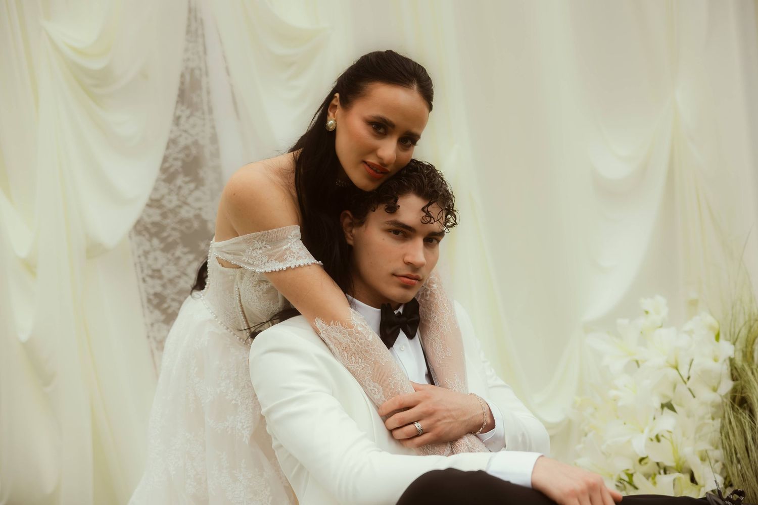 Bride in white off-shoulder gown embraces groom in white suit against elegant draped white curtain backdrop.