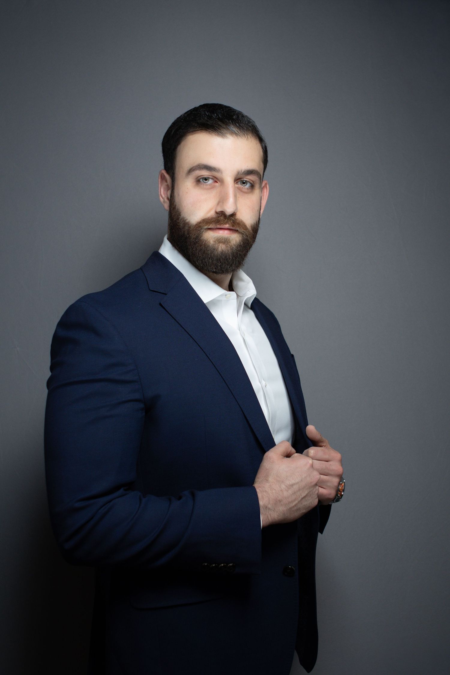 Bearded businessman in navy blue suit and white shirt posing confidently against gray background.